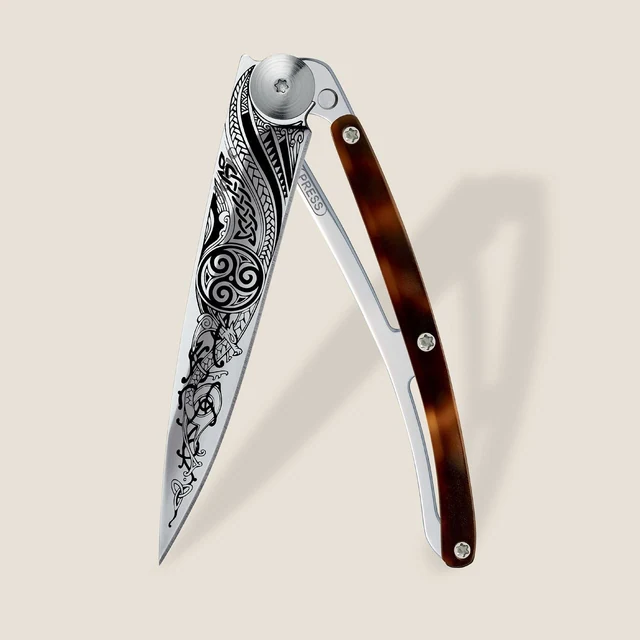 Complete Deejo Celtic folding knife with intricate blade artwork and an amber-patterned acetate handle on a warm neutral background