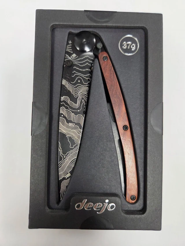 Complete Deejo Topography folding knife with contour-line blade design, serrated edge and coral-wood handle in its black presentation tray