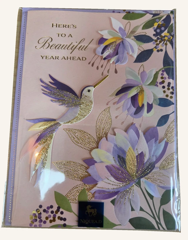 The actual hummingbird birthday card with layered purple flowers, golden details and a pink background in its original clear sleeve.